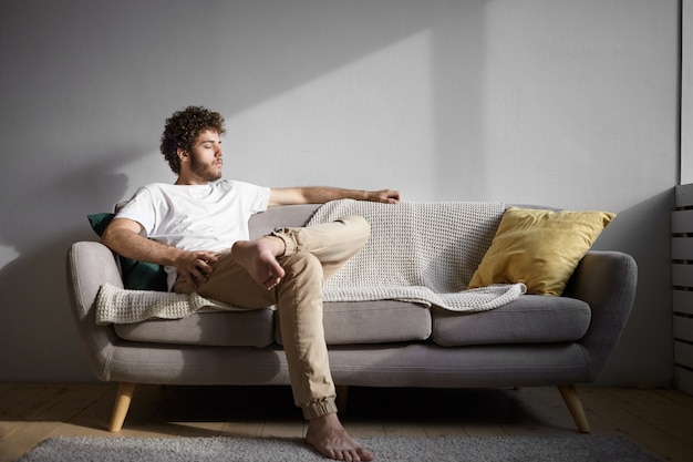 Free photo people, lifestyle, rest and relaxation concept. picture of handsome guy with bare feet resting indoors, sitting on cozy couch and closing eyes. stylish young man with stubble relaxing at home alone