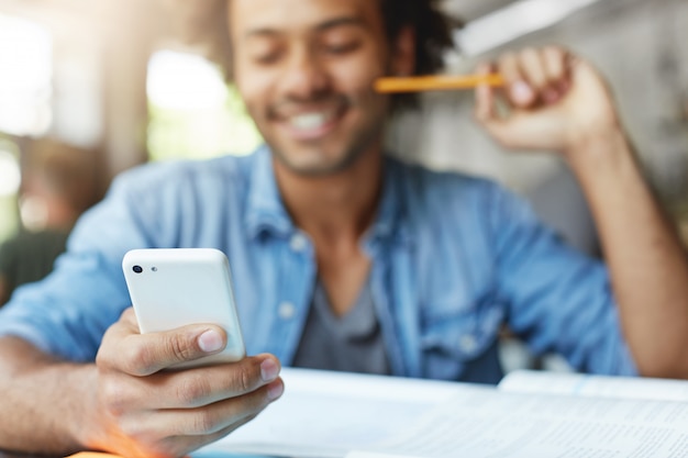 Free photo people, lifestyle, technology and communication concept. handsome bearded dark-skinned male student wearing blue shirt using cell phone, browsing newsfeed via social networks, laughing at memes