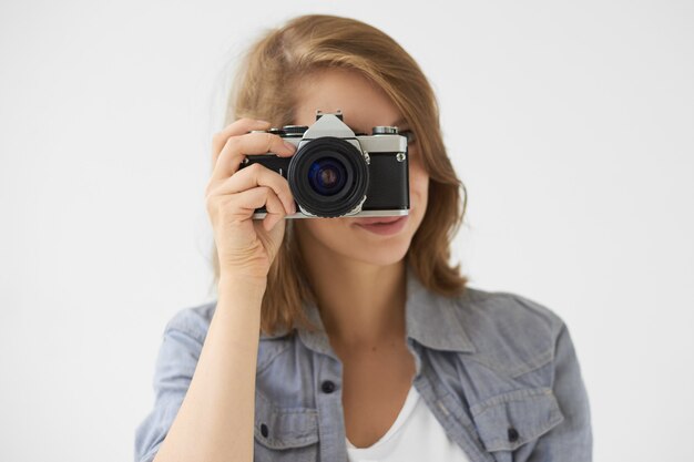 People, lifestyle and technology concept. Studio shot of stylish girl holding roll film camera at her face, taking picture of you. Young woman photographer using vintage device to take photo