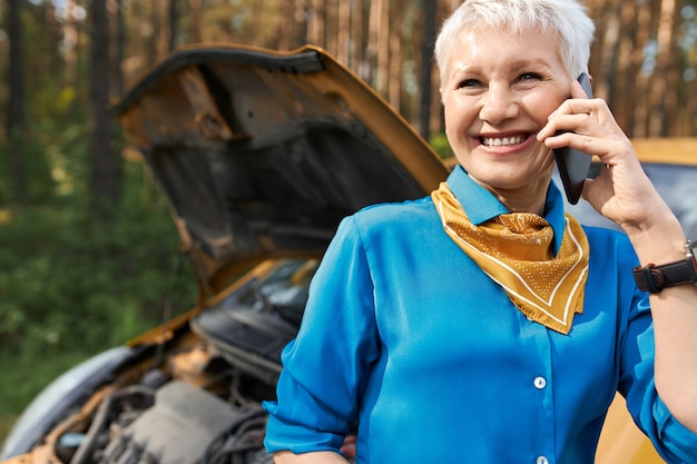 Free photo people, lifestyle, transportation and modern technology concept. beautiful blonde retired woman standing by broken car with open hood, calling roadside assistance, asking for help, smiling