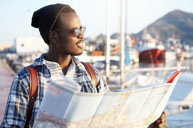 People, lifestyle, traveling and adventures concept. Man on quay surrounded by yachts and ships in fashionable hat and mirror sunglasses holding paper guide looking aside at sea with pleased smile