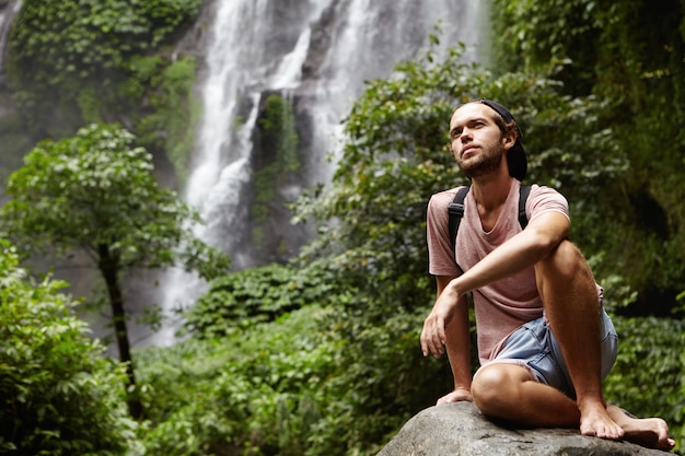 Free photo people, nature and adventure. young hipster with backpack sitting on big rock by waterfall