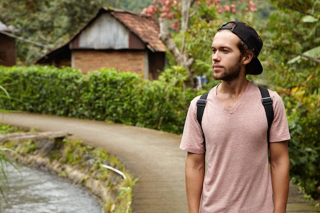 Free photo people, nature and summer concept. stylish young unshaven hipster man wearing snapback and backpack relaxing outdoors while walking along country road in rural area