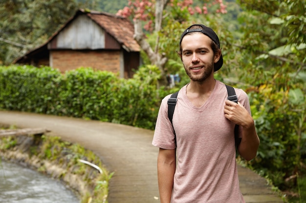 Free photo people, nature and summer concept. stylish young unshaven hipster man wearing snapback and backpack relaxing outdoors while walking along country road in rural area