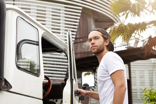 People, nature and transportation concept. Young hipster smiling happily, opening door of his white off-road vehicle