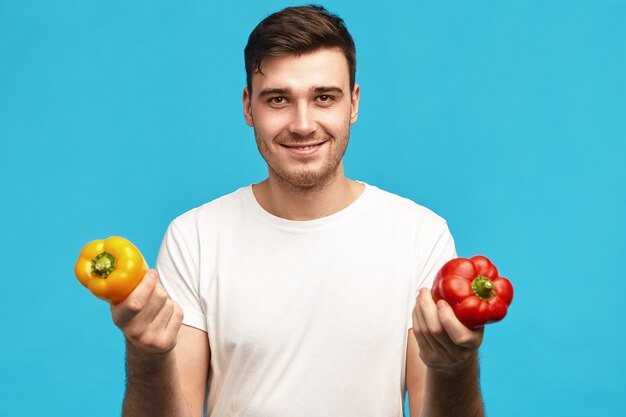 People, organic food, nutrition, vegetarianism and healthy lifestyle concept. Portrait of handsome positive young male wearing white t-shirt holding red and yellow bell peppers, going to make salad