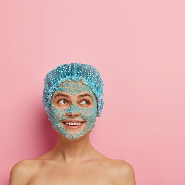 People, purity and facial treatment concept. Satisfied smiling young woman applies blue sea salt scrub, has naked body, clean glow skin, wears showercap, looks aside, has cosmetology procedure