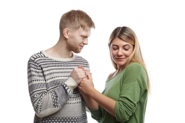 People, relationships and romance concept. Attractive young unshaven male confessing his love to pretty cute girl while holding both her hands, woman looking down with shy expression, blushing