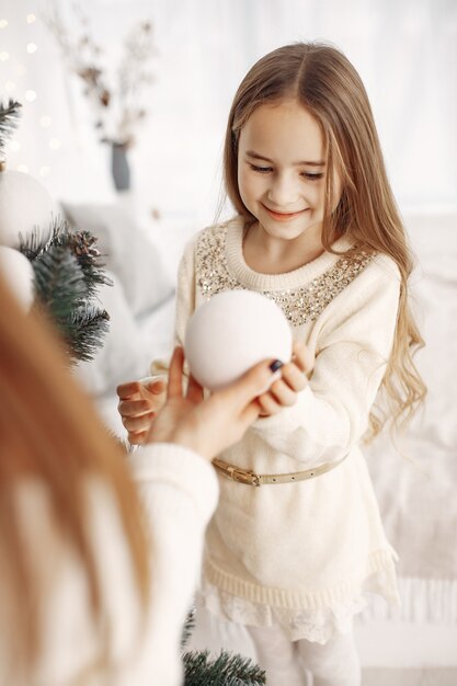 People reparing for Christmas. Mother playing with her daughter. Family decorated Christmass tree. Little girl in a white dress.