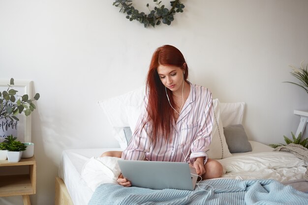 People, rest, relaxation and modern technology concept. Picture of beautiful redheadyoung female in striped pajamas sitting on undone bed using laptop and earphones while watching movie online