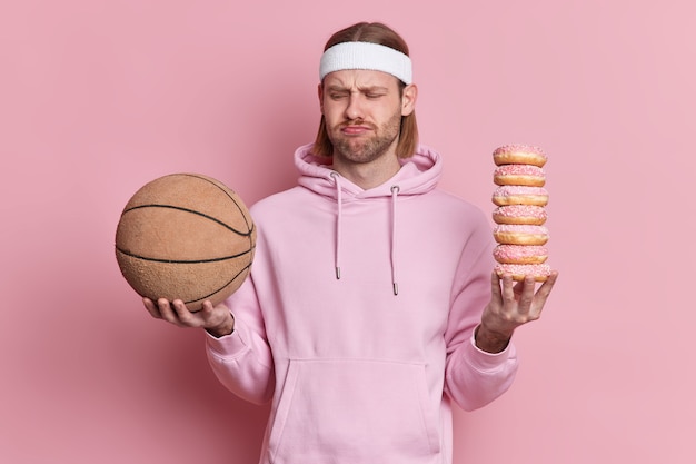 Free Photo people sport junk food concept. displeased sporty man looks unhappily at basketball ball holds pile of sweet appetizing donuts doesnt want to play dressed in hoodie.