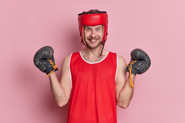 Free Photo people sport motivation concept. positive man has training in gym wears protective headgear boxing gloves t shirt has cheerful expression after winning sport game