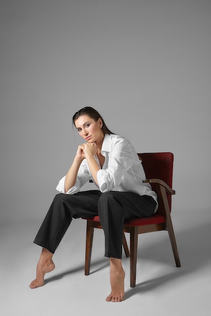 People, style, fashion, clothes and interior concept. Vertical portrait of stylish beautiful barefoot young female in men's formal clothing, sitting in chair like man with her legs wide apart