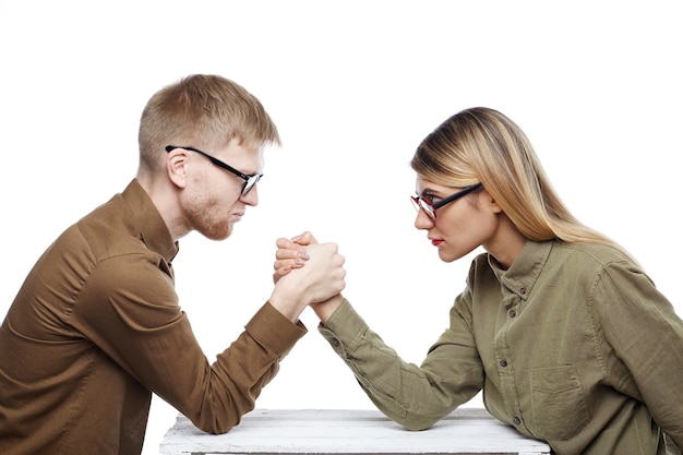 Free Photo people, teamwork, cooperation and competition concept. side view of young female and bearded male colleagues both wearing glasses arm wrestling, staring at each other with confident determined looks