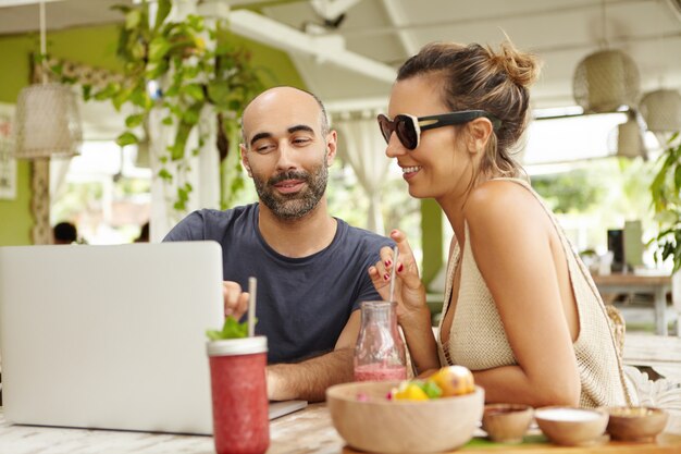 People, technology and communication. Adult couple using laptop computer at cafe, sitting at table with fresh drinks. Handsome man showing something to his girlfriend on notebook.