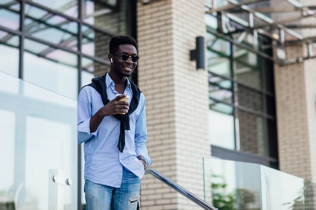 People, technology and lifestyle - happy young man with headphones and smartphone listening to music in city with cup of coffe.