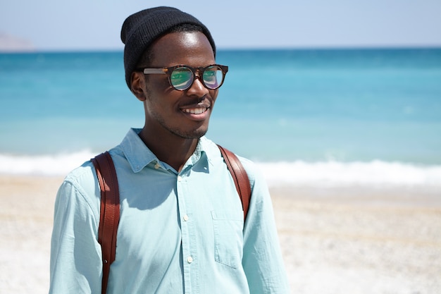 People, travel, holidays and tourism concept. Joyful young dark-skinned male backpacker enjoying picturesque view and seascape during summer vacations in resort town, walking on beach alone