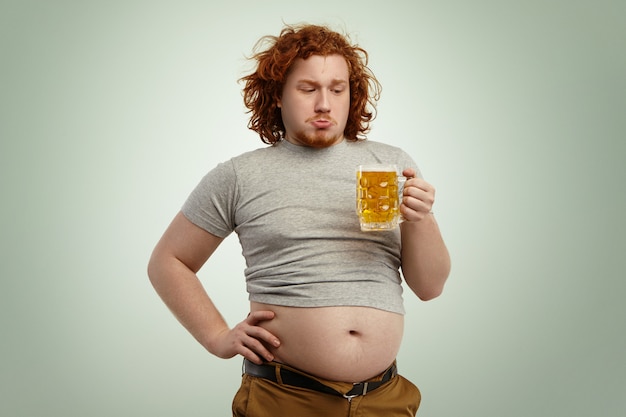 Free photo people, unhealthy lifestyle, obesity and gluttony. overweight fat young european man with curly red hair holding glass of beer, feeling hesitated deciding whether to drink it or not after good dinner