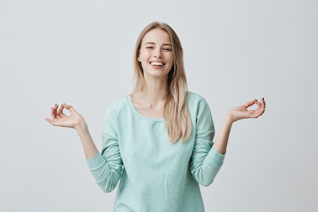 People, yoga and healthy lifestyle. Caucasian young blonde woman keeping eyes closed while meditating indoors, practicing piece of mind, keeping fingers in mudra gesture, smiling broadly