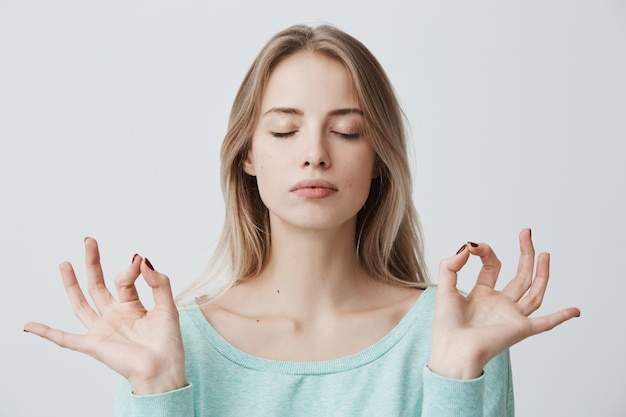 Free photo people, yoga and healthy lifestyle. gorgeous young blonde woman dressed in light blue sweater keeping eyes closed while meditating indoors, practicing peace of mind, keeping fingers in mudra gesture