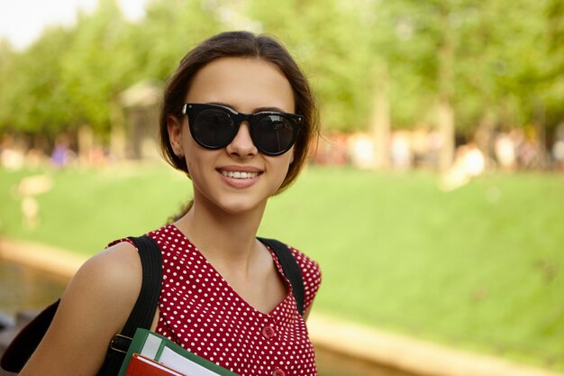 People, youth, school and education concept. Fashionable happy positive college girl wearing black shades and carrying backpack enjoying nice summer weather, going home from classes,