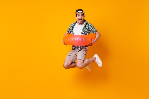 Perky man in shorts and shirt put on inflatable circle and jumps on orange space.