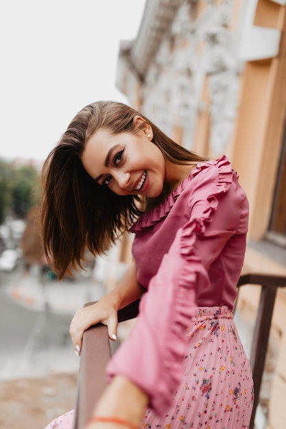 Perky woman in soft tender blouse looks at camera coquettishly Portrait of stylish European woman stretching towards camera on balcony