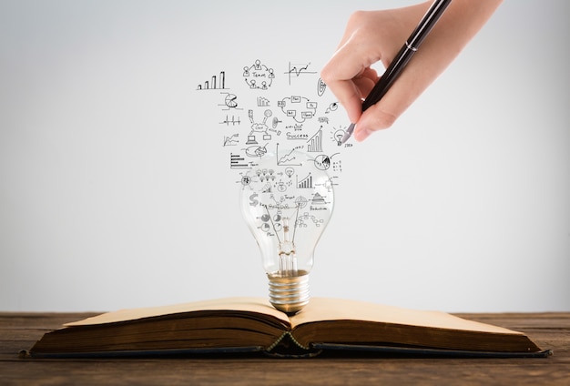 Person drawing symbols coming out of a light bulb on top of a book