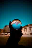 Free photo a person holding a glass ball with the reflection of buildings and the blue sky