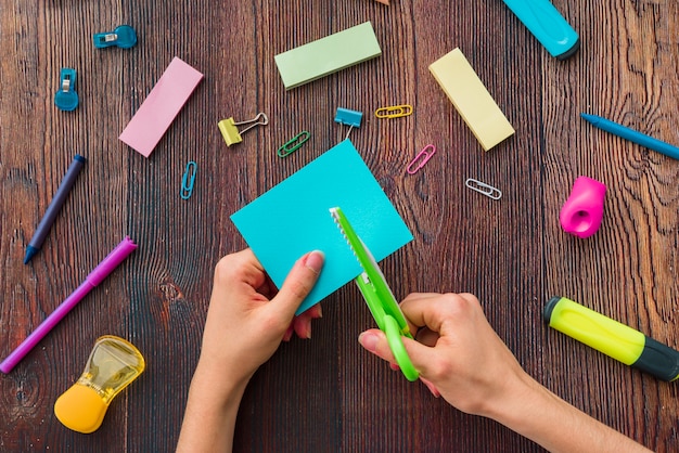 Person's hand cutting blue paper over the school accessories on wooden table