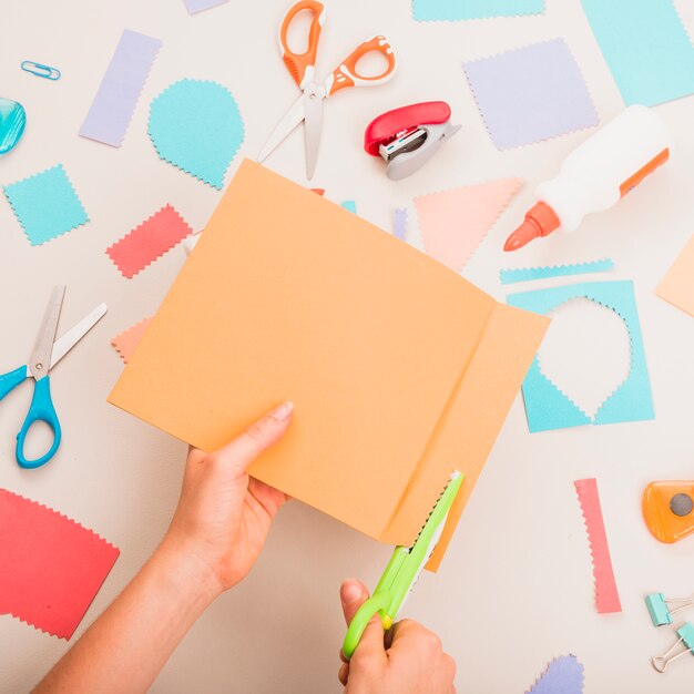 Person's hand cutting colorful paper over school supplies on table