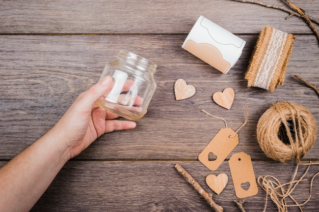 Free photo a person's hand holding an empty glass bottle with lace ribbon; wooden heart shape; tags and spool thread on table