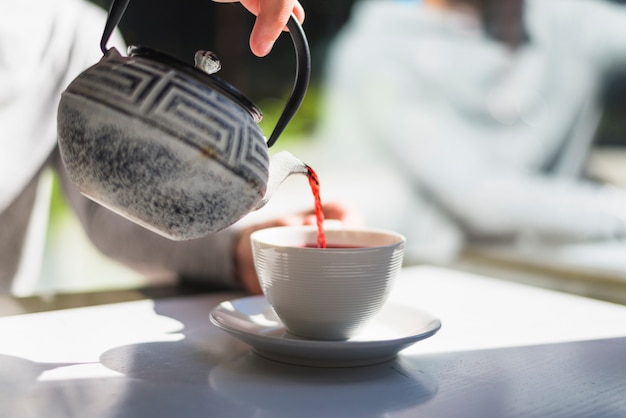 A person's hand pouring red tea in the white ceramic cup on the white table in the sunlight