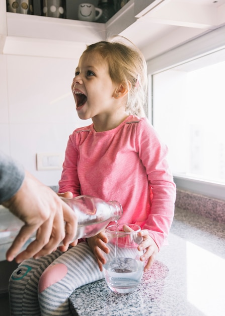 A person's hand pouring water in glass while girl sitting on kitchen counter