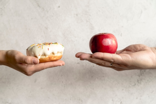 Free photo person's hand showing donut and apple in front of concrete background