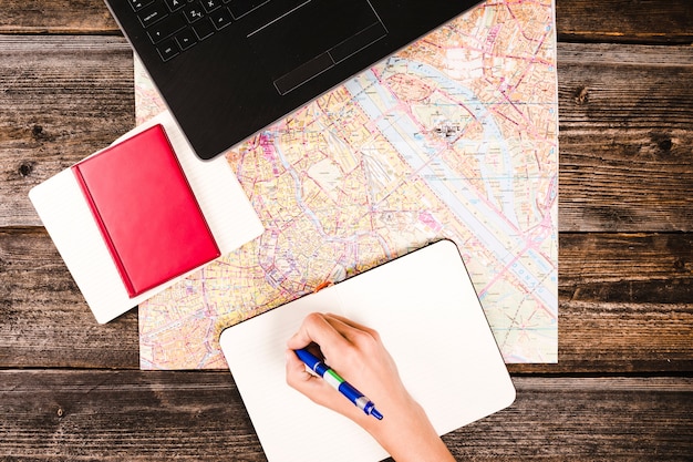 Free Photo person's hand writing on notepad with diary and map on wooden table