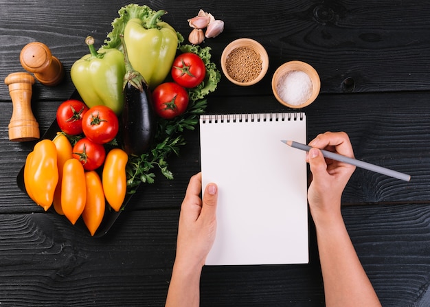 Free photo person's hand writing on spiral notepad near vegetables and spices on black wooden surface