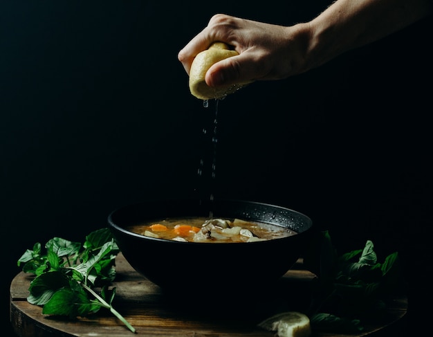 Person squeezing lemon on soup in a black bowl with a dark wall