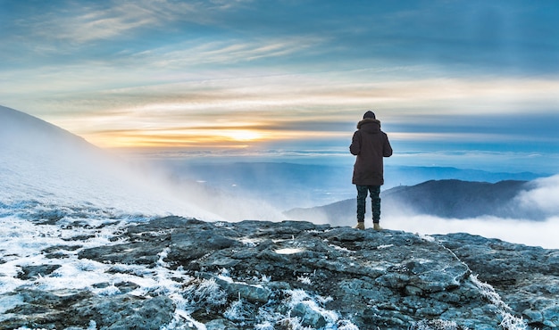 Person standing on a snow covered cliff over the breathtaking view of mountains under the sunset