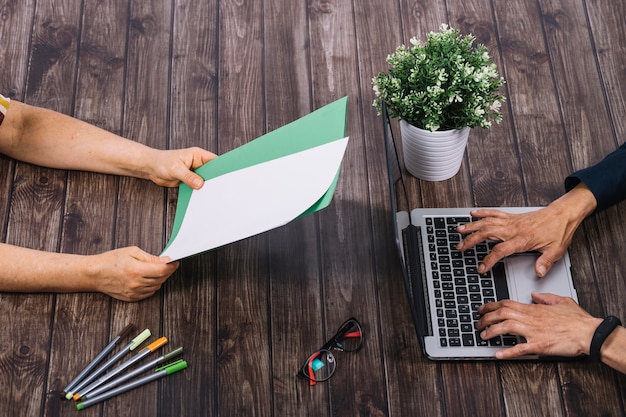 Free Photo a person typing on laptop with his colleague holding blank white and green blank paper on wooden table