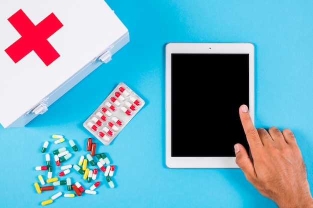 Free Photo a person using digital tablet with first aid kit and capsules on blue background