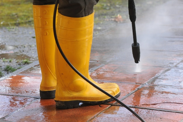 Person wearing yellow rubber boots with high-pressure water nozzle cleaning the dirt in the tiles