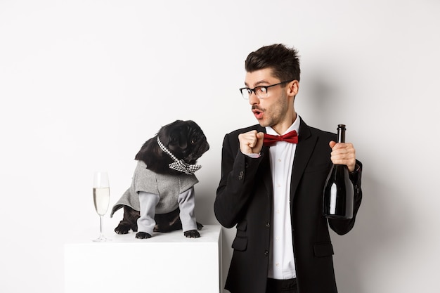 Pets, winter holidays and New Year concept. Happy young man celebrating Christmas with cute black dog wearing party costume, puppy looking at owner, white background.