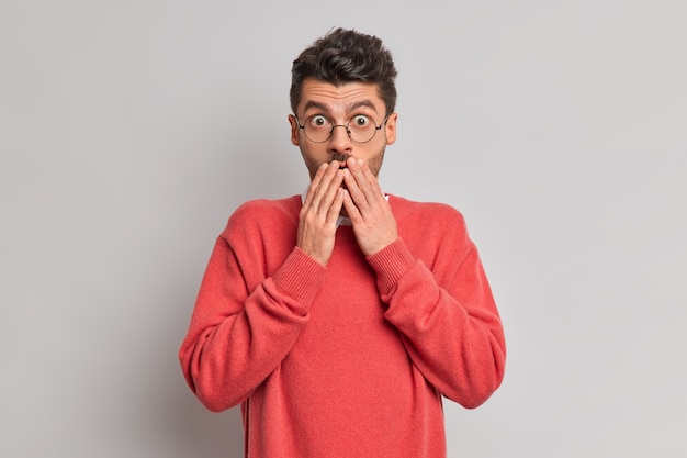 Free photo photo of amazed young european man keeps hands on mouth stares shocked at camera reacts on unexpected relevation