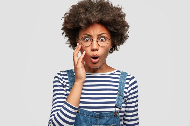 Photo of anxious African American female looks surprisingly and raises eyebrows in bewilderment