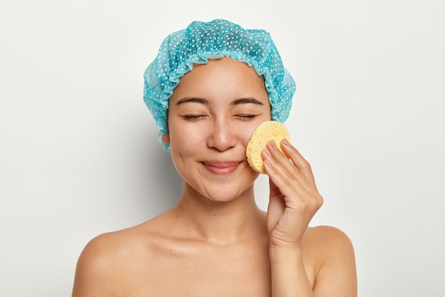 Photo of Asian woman cleans face with cellulose sponge, cares about skin, has cosmetology treatments, eyes closed, stands with bare body, stands against white wall. Spa procedures, makeup removal