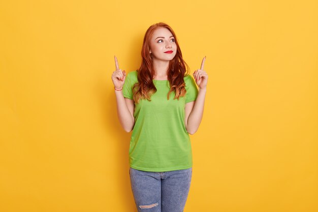 Photo of attractive dark haired young European woman pointing with index fingers up at copy space, poses against yellow space