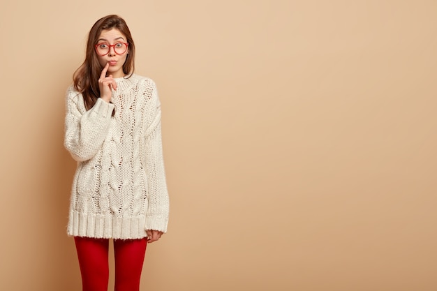 Free photo photo of attractive woman keeps index finger near folded lips, looks curiously, has surprised expression, listens intriguing information, wears casual winter sweater, stands over beige wall