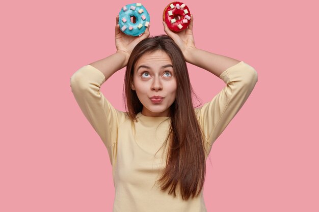 Photo of attractive woman with dark hair, keeps colourful doughnuts over head, demonstrates junk food