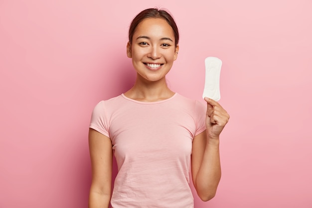 Free Photo photo of attractive young lady with asian appearance, holds clean sanitary napkin, satisfied with its quality, uses intimate product during menstruation or periods, isolated on pink wall